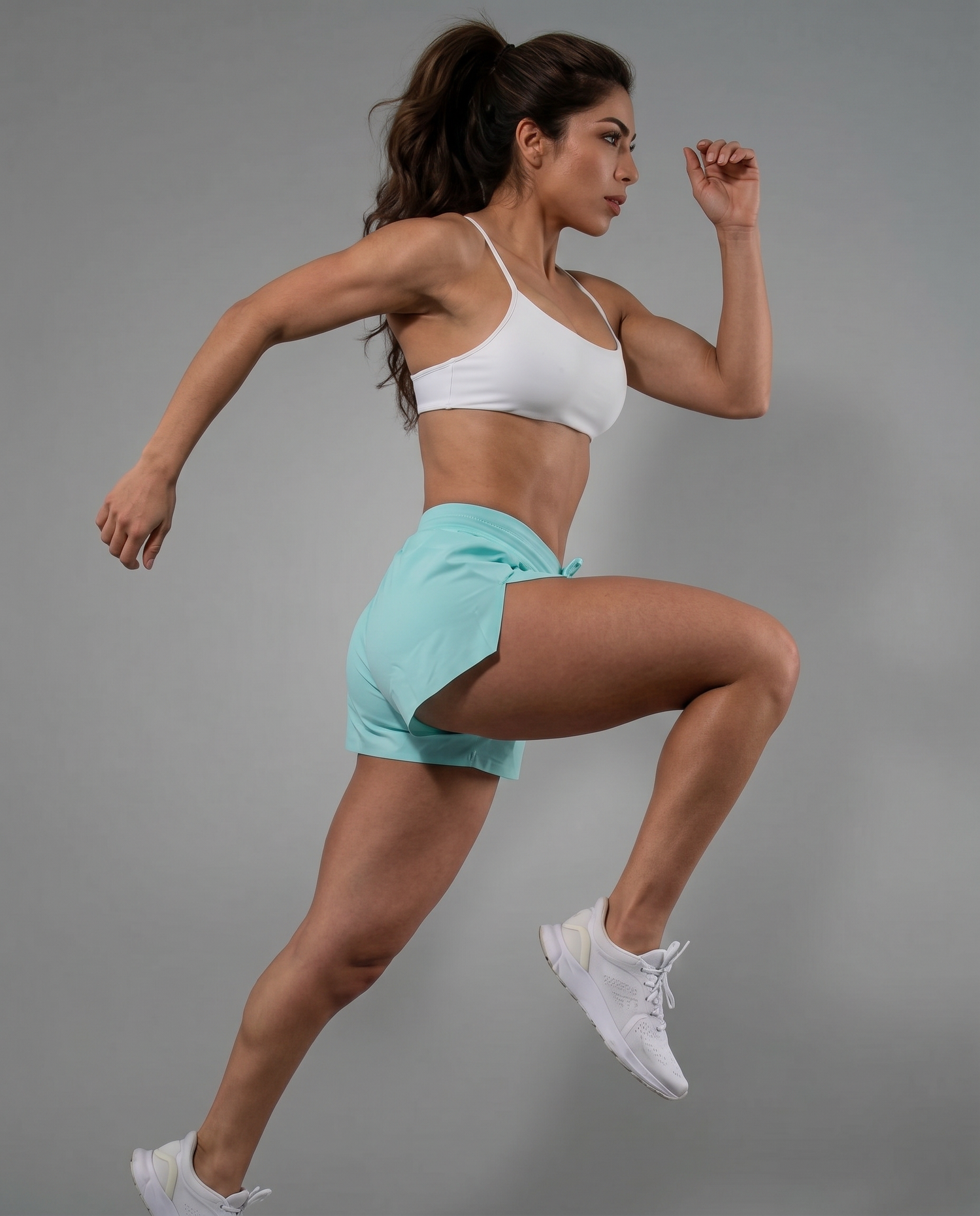 Woman running mid-stride in mint green high-waisted booty shorts and white sports bra against a gray studio backdrop, Gym Crush collection by Violate the Dress Code